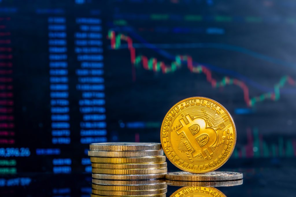 A stark of cryptocurrencies placed in front of a trading screen. Close-up of golden Bitcoins on a dark reflective surface and the histogram of decreasing crypto in the background