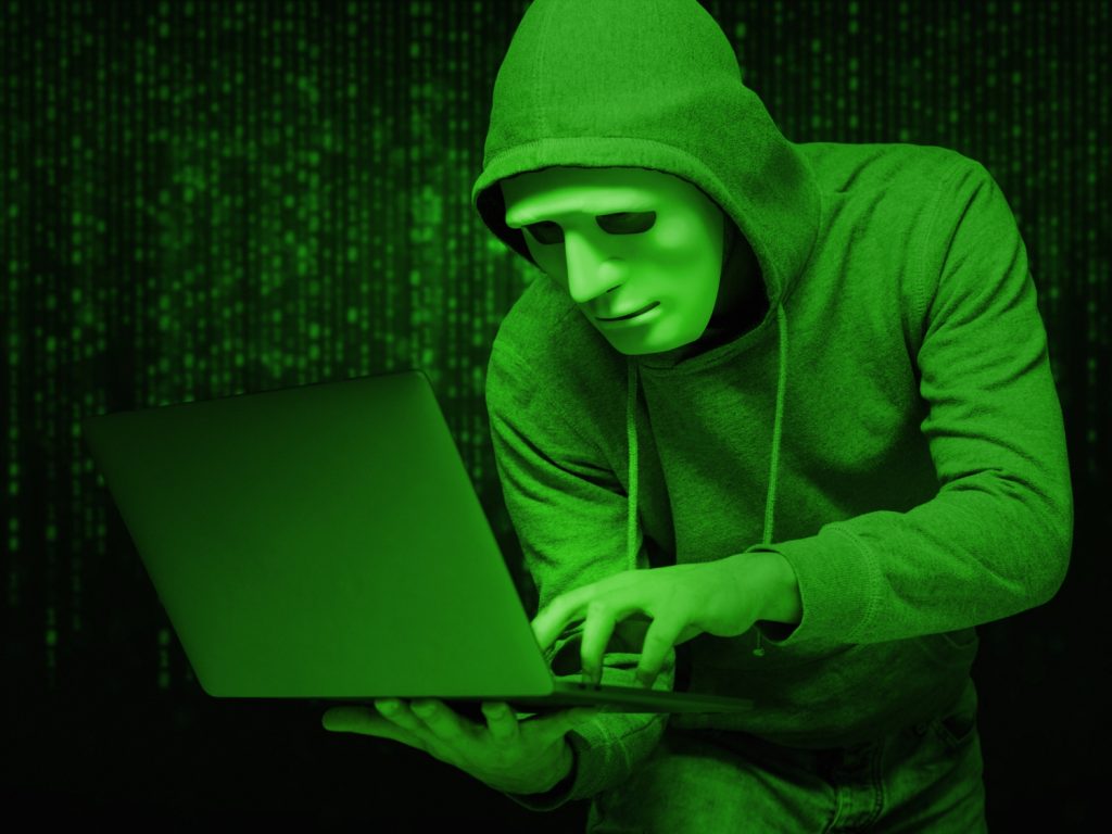 A picture of a crypto hacker weaking a mask while holding a laptop