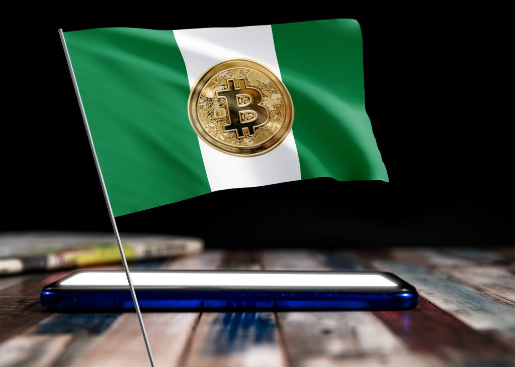 A Nigerian flag featuring a Bitcoin symbol waves above a smartphone on a wooden surface, symbolizing Nigeria’s evolving stance on cryptocurrency regulations in 2025.