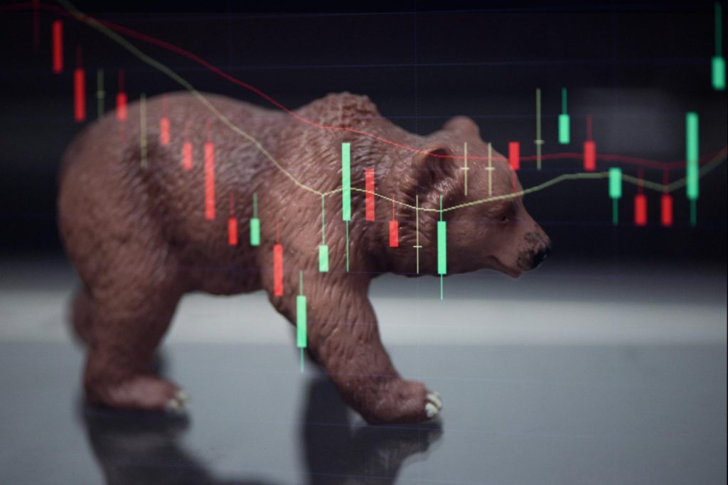 A toy bear walking across a screen showing red and green candlestick crypto charts, symbolizing a bear market