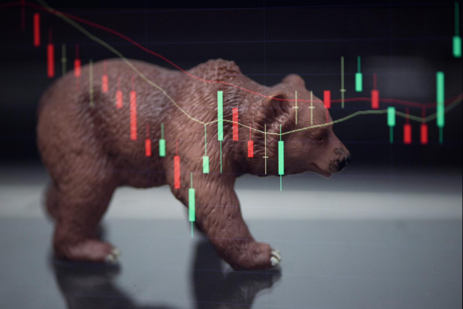 A toy bear walking across a screen showing red and green candlestick crypto charts, symbolizing a bear market