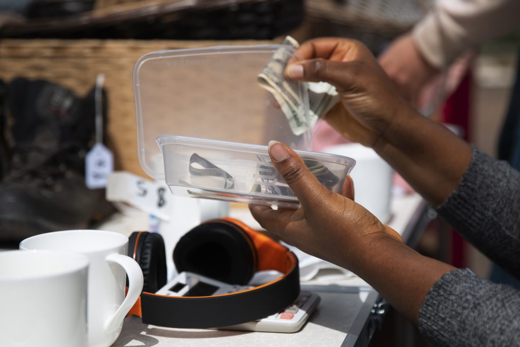 A person counting cash and organizing small electronics and accessories at a garage sale, symbolizing the risks of handing out money without verifying legitimacy—relatable to online crypto scams.