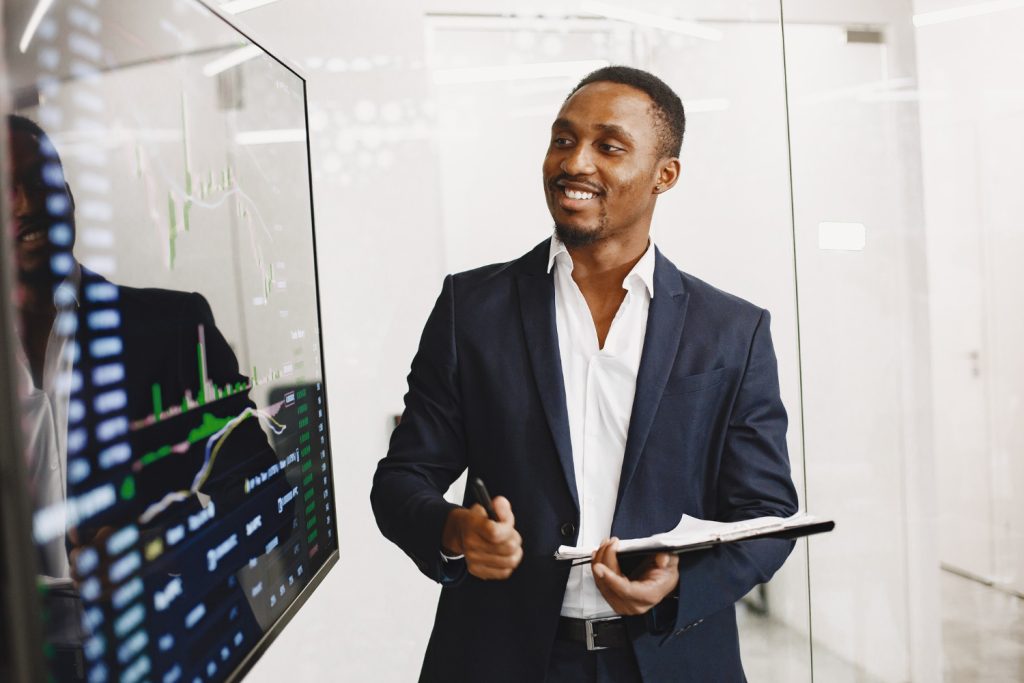 A confident African man in a suit giving a presentation on cryptocurrency trading charts in a modern office setting.