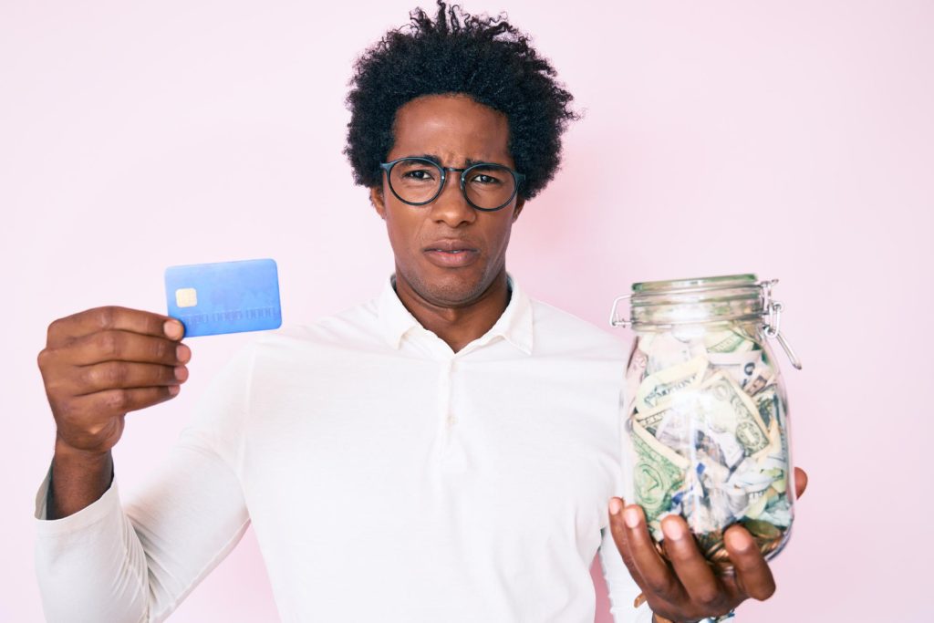 Confused African-American man with afro hair holding a credit card in one hand and a jar of cash in the other, expressing doubt or uncertainty about financial decisions.