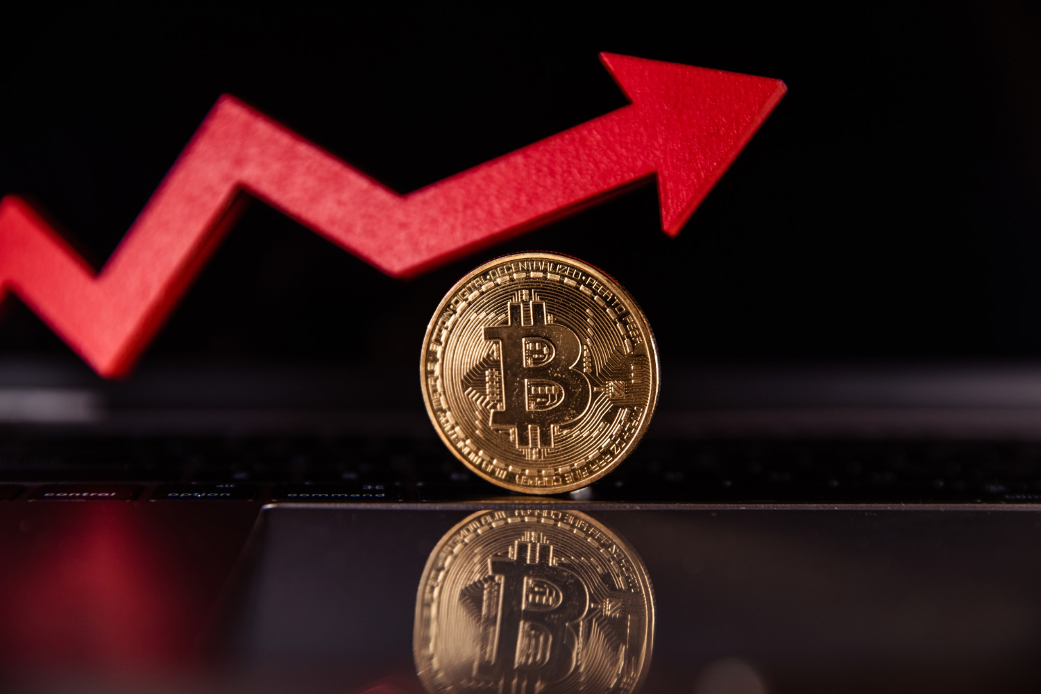 A shiny gold Bitcoin coin standing upright on a reflective surface, with a bold red upward arrow in the background indicating a price surge.