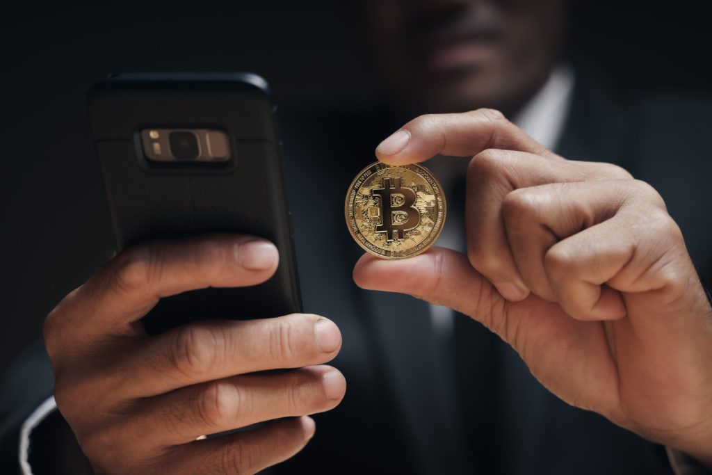 Close-up of a person in a suit holding a gold Bitcoin coin in one hand and a smartphone in the other.