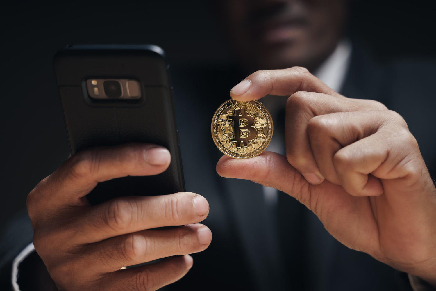 Close-up of a person in a suit holding a gold Bitcoin coin in one hand and a smartphone in the other.