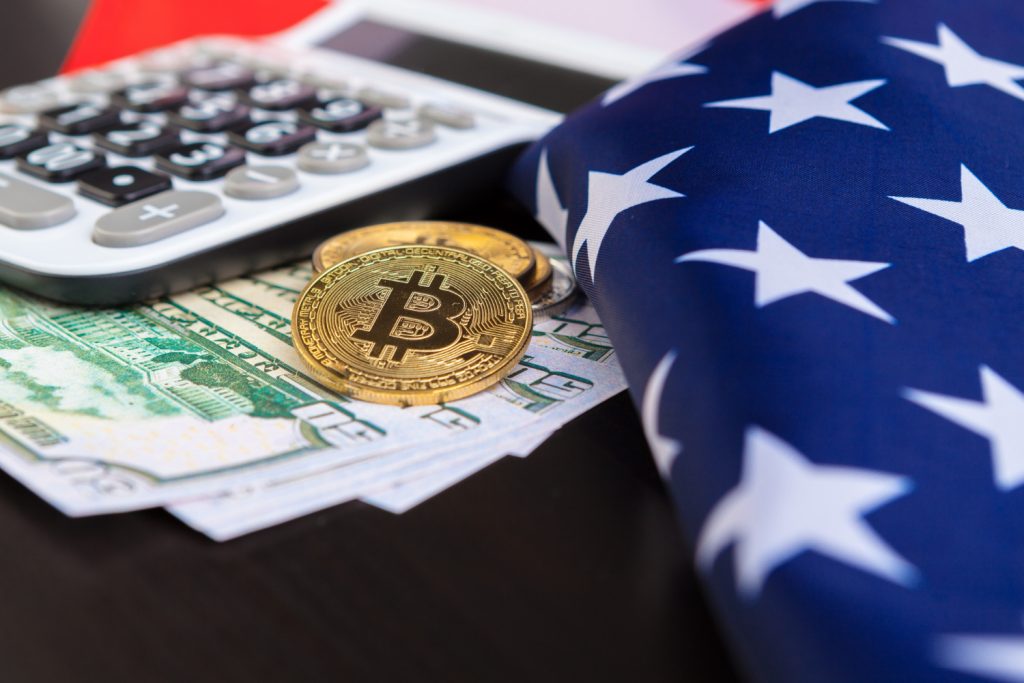 Close-up of physical Bitcoin coins placed on US dollar bills, alongside a calculator and partially draped American flag, symbolizing the intersection of cryptocurrency and US financial policy.