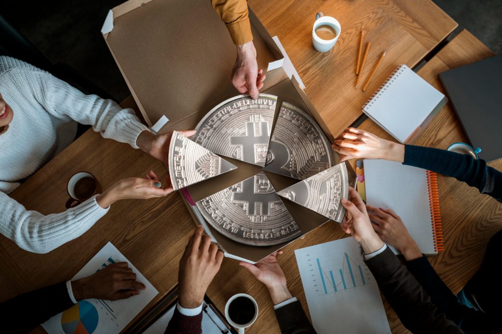 “Top view of people around a table sharing slices of a large Bitcoin-shaped pizza, symbolizing cryptocurrency investment and distribution.”
