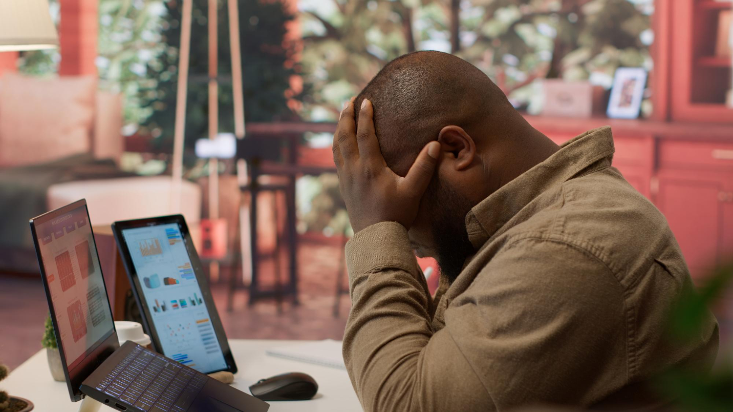 A frustrated man sitting at his desk with his hands on his head while looking at multiple computer screens displaying financial or cryptocurrency charts.
