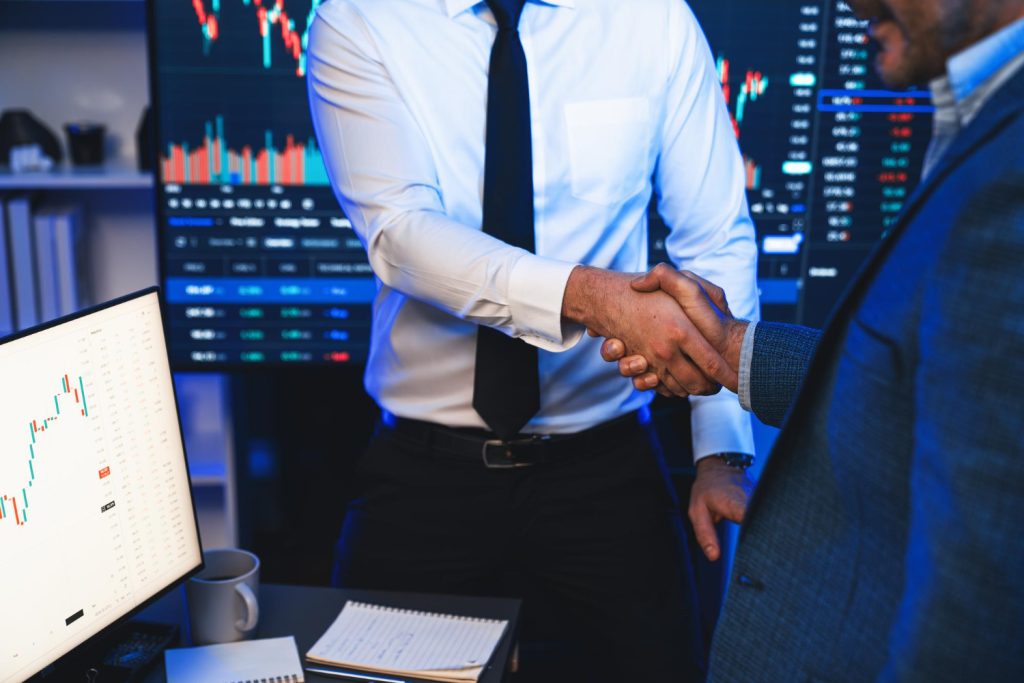 Two professionals shaking hands in front of trading screens displaying financial charts and market data.”